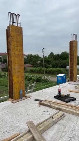 The video shows a construction site with two large concrete columns being formed. The columns are made of yellow and brown plywood forms, with rebar sticking out of the top. The ground is covered with concrete, and there are various construction materials scattered around, including wooden planks, a ladder, a toolbox, and a portable toilet. The sky is cloudy. The video pans across the scene, showing the columns from different angles. There are no people visible in the video.