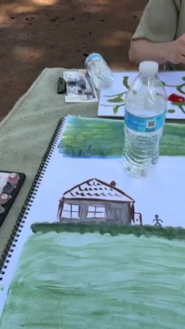 The video begins with a close-up of a painting on a table, featuring a house and green fields. A bottle of water is placed on top of the painting. The camera then pans to show a young man with curly hair, wearing a light green t-shirt, painting on a separate canvas. He is holding a paintbrush and appears to be adding detail to a painting of red mushrooms with green leaves. Several small bottles of paint are arranged on the table, along with pencils. In the background, there are picnic tables and trees, suggesting an outdoor setting, possibly a park or campsite. Further in the background, other people can be seen sitting at picnic tables and under a canopy. The video then shifts focus to another person, wearing a green hat, who is painting on a canvas propped on an easel. This painting is abstract, with a mix of red and gray colors. A smartphone, more paint bottles, and other art supplies are visible on the table in front of this person. The scene continues to show the two individuals engaged in painting outdoors, surrounded by nature.