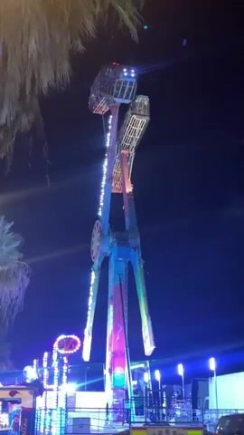 This is a video of an amusement park ride at night. The ride appears to be a pendulum-style thrill ride with multiple arms that swing and spin. The ride is brightly lit with colorful LED lights, creating a festive atmosphere. The sky is dark, indicating it is nighttime. Palm trees are visible in the background, suggesting a warm climate, and the location is identified as Canet de Mar, Spain. The ride is in motion, with parts of it swinging high into the air. There is a sense of excitement and thrill associated with the activity of riding such a machine at an amusement park. The lighting and motion of the ride suggest it is a popular attraction. The video captures the vibrant energy of the amusement park at night.