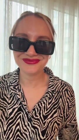 A woman in a zebra-striped shirt and sunglasses stands in front of a window with sheer curtains. She has her blonde hair pulled back in a bun. She crosses her arms, turns her head slightly, and looks around. Buildings are visible outside the window.