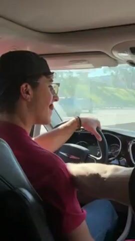 A young man is driving a car. He wears a dark baseball cap, glasses, a maroon shirt, and a bracelet on his wrist. He glances to his right as he holds the steering wheel, waiting in traffic. Cars are visible outside the windshield. The interior of the car is light-colored. The video is shot from the passenger seat, offering a clear view of the driver.