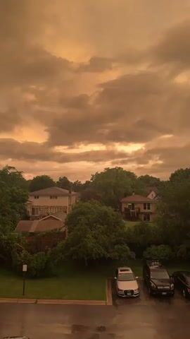 The video shows a view of houses and trees under a cloudy sky. The sky is a predominant orange-brown color, suggesting an unusual atmospheric condition like smoke or dust. In the foreground, there is a parking lot with several cars, and some wet pavement, indicating it may have recently rained or is still raining. The houses are suburban in appearance, with some green trees surrounding them. The overall impression is one of a somewhat somber or dramatic atmosphere due to the sky's color.