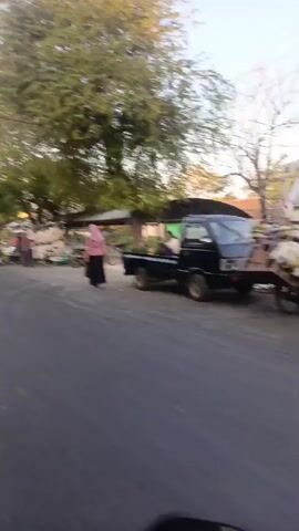 The video shows a street scene with several people and vehicles. On the right side of the road, a black pickup truck is parked, and two people are standing near it. There are also some plants or goods being transported. In the middle of the scene, a woman in a pink hijab is walking along the roadside. Further down the street, there is a line of motorcycles parked, and several people are gathered around them, possibly vendors or customers. Some of the people appear to be interacting with each other. On the left side of the road, more motorcycles and a white truck are visible. The overall environment appears to be an outdoor market or a busy street with vendors. The lighting suggests it might be late afternoon or early morning.
