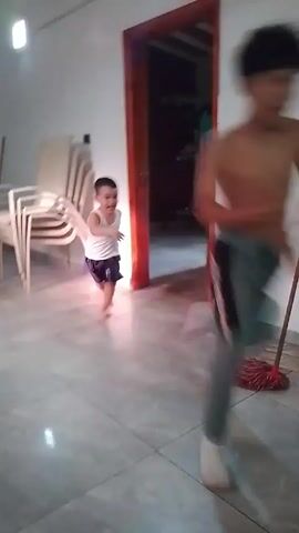A brief video depicts a child in a white tank top and dark shorts running barefoot through a room with tiled floors. The room contains a mop and bucket in the foreground, as well as a dining area with a glass-topped table and black chairs. Further back, there is a wooden cabinet, a bicycle, and a scooter. A shirtless person is briefly visible in the doorway of another room.  The camera is relatively still but moves slightly to follow the child. There is no discernible audio in the clip.