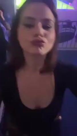 A woman with dark hair is recording a selfie video at what appears to be a club. She is wearing a black shirt. Behind her, a man with a mustache can be seen occasionally moving in and out of focus. The woman is sticking out her tongue and smiling, seemingly enjoying the music or atmosphere around her. The lighting is dim with some blue accents, and the background is blurry, suggesting movement or a lively environment. Heavy beats can be heard in the video.