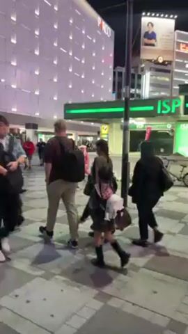 The video shows a crowded urban street at night. People are walking in various directions across a crosswalk. The buildings are lit up, some with digital displays. One person is wearing an orange costume. The camera moves erratically, suggesting it is held by someone walking through the crowd.