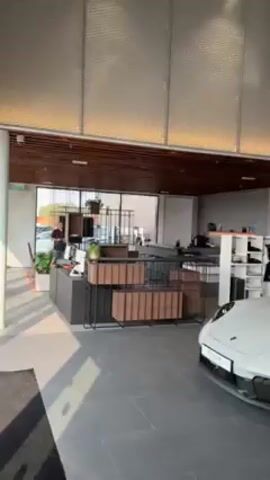 The video shows the interior of a Porsche dealership. The space is well-lit with modern architectural elements. Two Porsche vehicles are prominently displayed, one white sports car and a light gray SUV. A small toy drone sits on the hood of the SUV. The dealership features a reception area with a desk, shelving, and seating. Large windows offer an outside view, and the brand name “PORSCHE” is visible in bold, capitalized letters above a vehicle.