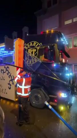 The video shows a nighttime street scene with a parade float. The float consists of a dark blue truck with a platform built on the back.  The truck has a large gold-colored emblem with the letters "RT HABIS" on the side. It is decorated with Christmas lights. On the back platform, two people are visible, possibly DJs or performers. A man in an orange and white safety jacket stands near the truck, holding a blue hose.  A small crowd of people is gathered around the truck, including a person wearing a pink knit hat. The side of the float has white panels with blue decoration and writing, including "1979 e.V." and the name "Prinzesin MIRI".  The street appears wet, reflecting the lights. The lighting is provided by the truck, streetlights, and the float decorations. The overall atmosphere seems festive and lively.