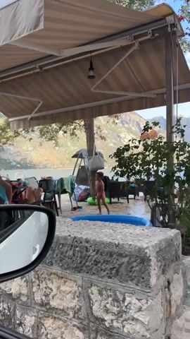 A wide shot shows a shaded outdoor seating area with a stone wall in the foreground. A person is lying on a chair under an umbrella, partially covered by a towel with a cartoon character on it. A child in swimwear stands nearby, and a blue inflatable pool is on the ground. The background features a body of water with mountains in the distance. The camera pans to the left, showing more of the seating area with tables, chairs, and umbrellas. A woman and two children are visible in the water. The camera continues to pan left, showing more of the water and distant mountains. The video ends with a closer view of the water with people swimming and mountains in the background.