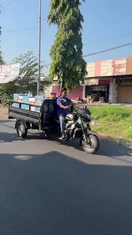 A person is riding a three-wheeled vehicle with a cargo bed down a paved road. The vehicle is black and appears to be a motorcycle with a trailer. The cargo bed is filled with boxes. The person is wearing a blue t-shirt and jeans. The road is lined with buildings and trees. The sky is clear and blue.