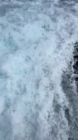 The video captures a dynamic scene of a boat's wake in the ocean. Initially, the camera focuses on the churning white foam and water close to the boat, showcasing the turbulent effect of its passage. As the video progresses, the camera pulls back, revealing a wider expanse of the ocean with the boat's wake stretching out behind it. In the distance, a coastline is visible, dotted with buildings and covered by a hill. The sky above is a clear blue. The overall impression is of a boat moving through the sea, leaving a distinct trail on the water's surface, with a picturesque coastal town in the background under a bright, sunny sky.