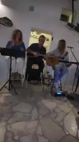 The video shows a group of people at an outdoor restaurant. A woman is sitting at a table with a checkered blue and white tablecloth. Two men are sitting on stools playing guitars and singing into a microphone. Other people are seated at tables, and some are walking around. The restaurant has a white exterior with blue accents. The floor is made of stone tiles. There are string lights hanging overhead.