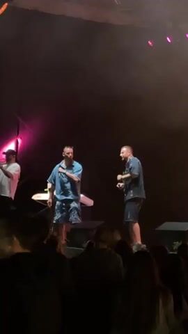 Three men are performing on a dark stage illuminated by colorful stage lights. The man on the left wears a white short-sleeved shirt and stands near a table with electronic equipment, holding a microphone. The man in the center wears a light blue short-sleeved shirt and patterned shorts, holding a microphone and gesturing. The man on the right wears a dark patterned short-sleeved shirt and dark shorts, holding a microphone and moving to the music. A crowd of people, mostly obscured by darkness, is visible in the foreground, facing the stage. The stage lights transition between yellow and pink hues.