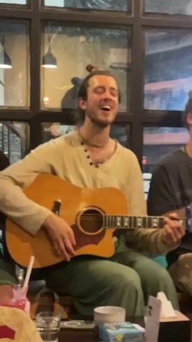 A young man is seated indoors, enthusiastically playing an acoustic guitar and singing. He has a beard, long dark hair tied in a bun, and is wearing a light beige, long-sleeved tunic with a V-neck and dark buttons, and green pants. A simple necklace is visible around his neck. His eyes are closed, and he has a wide smile on his face, indicating joy and immersion in the music.

The light-colored guitar has a dark pickguard, and a capo is clamped on its neck. In front of him, on a table, are several items: a glass containing a pink drink with a straw, a clear glass of water, a small white bowl, a blue box with the word "SACRED" printed on it, and a small white paper dispenser.

The background consists of a large window divided into multiple panes by dark frames. Reflections of indoor lights and dimly visible figures can be seen in the glass, suggesting it is evening or night. Another person is partially visible on the right side of the frame, seated next to the guitarist. The setting appears to be a cozy cafe or gathering space in Pokhara, Nepal.