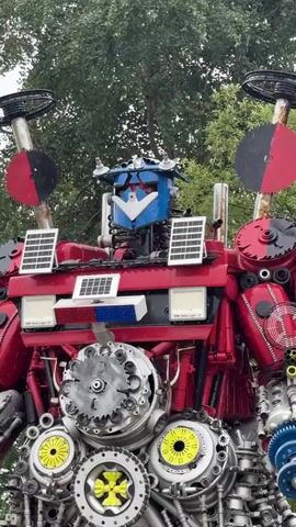 This video shows a large, red, and blue robot sculpture made of recycled metal parts. The robot is standing in an outdoor setting with trees in the background. The robot has solar panels on its chest, and its head is blue with a white faceplate and red accents. The robot's body is made of various metal components, including gears, pipes, and car parts. The robot's arms are extended outwards, and its legs are bent as if it is about to move. The video is filmed from a low angle, looking up at the robot, which gives it a towering and imposing presence. The lighting is somewhat dim, suggesting it might be an overcast day. The robot appears to be a static sculpture, not in motion.