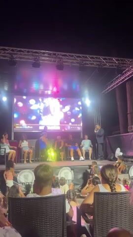 An outdoor stage at night features a large screen displaying a colorful, abstract pattern. On stage, a man in a dark suit and tie stands holding a microphone, addressing an audience. In front of him, five people are seated in chairs. The man appears to be interacting with these individuals, possibly as part of a performance or presentation. The audience is seated in front of the stage, and the lighting suggests an evening event.