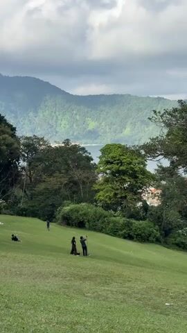 The video shows a landscape view with a large green hill in the foreground. Several people are scattered across the hill. In the middle of the hill, a couple is standing together, and the person on the right is holding up a phone, possibly taking a selfie. Further up the hill, another person is walking. To the left of the frame, a person is lying down on the grass. In the background, there is a dense forest, a body of water, and a mountain range under a cloudy sky. The overall scene suggests a leisurely outdoor setting.