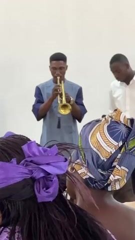A medium close-up shot of a man playing a trumpet while standing in front of a white wall. He is wearing a blue and gray shirt. Another man in a white shirt is standing to his right. In the foreground, the back of a woman's head wearing a blue headscarf is visible. The camera pans right, revealing a group of women sitting in a room. Many of them are wearing headscarves. The room has red and orange striped curtains.