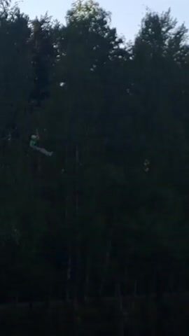 The video shows a person on a zipline over water. The person is wearing a green shirt, and a helmet, and their legs are straight. They are moving from left to right in the frame. Behind the person is a line of trees. The sky is blue.