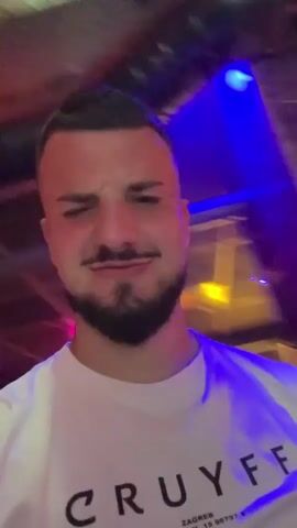 The video shows a man in a white t-shirt winking and sticking out his tongue. He has a beard and short hair. He is in a dimly lit environment with colorful lights, including blue and red. In the background, another person can be seen. The man then smiles and winks again. In the next scene, two men are visible, both smiling. The man in the foreground is wearing a white t-shirt with text on it. The person next to him is wearing a tan hoodie. There are also red lights visible in the background. The video continues with shots of the two men laughing and interacting. The lighting remains dim and colorful.