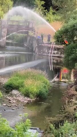 Firefighters are using hoses to spray water onto the remains of a bridge over a river. There are several firefighters visible, some wearing high-visibility vests, and one appears to be operating a ladder. The bridge structure is made of concrete and metal, and it looks old and damaged. The river is flowing and has some vegetation growing in it. The area is surrounded by trees. The water spray from the hoses is creating a mist that obscures some of the scene.