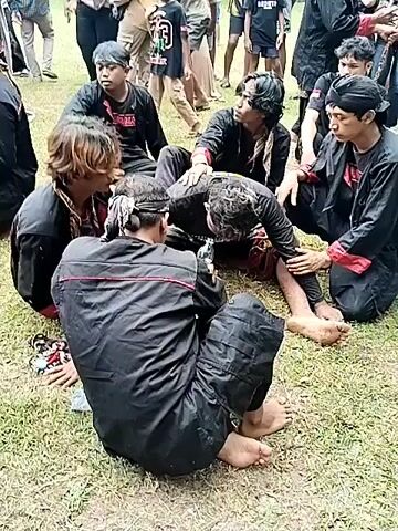 The video shows a group of people, mostly men, in a grassy outdoor setting. Several individuals are wearing black outfits with red and gold trim, some also have headbands. They are gathered around a man who is lying on the ground. He is being helped to drink water from a plastic bottle. Other people, including children, are standing around watching. A man with a black shirt that has the word "Kobestar" embroidered on the back is visible. The environment appears to be a public gathering or event.