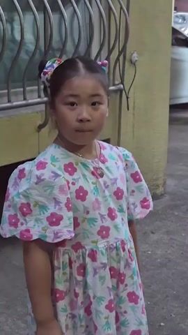 A young girl with two pigtails and a floral dress stands in front of a building. She is looking at the camera with a neutral expression. The background includes a metal fence and a yellow wall. The lighting appears to be dim, suggesting it might be evening or indoors.