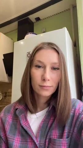A woman with shoulder-length brown hair, wearing a pink and gray plaid shirt over a white t-shirt, is filming herself from the chest up in what appears to be a kitchen. She is standing in front of a white refrigerator and a portion of a green wall. She begins by waving at the camera, then blows a kiss, covering her mouth with her hands. Next, she lowers her hands and opens her mouth, as if speaking. Finally, she brings her hands together in front of her face, holding a small, clear object between her thumb and index fingers, which she shows to the camera.