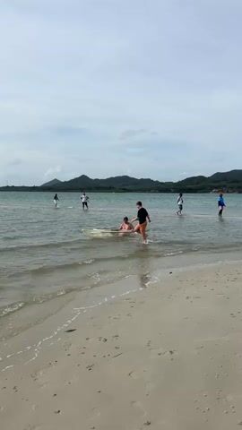 A group of people are at the beach. Two people are in a small boat in the shallow water. One person is pushing the boat while the other person sits in it. Several other people are standing in the water or on the beach in the background. The sky is cloudy and there are mountains in the distance.