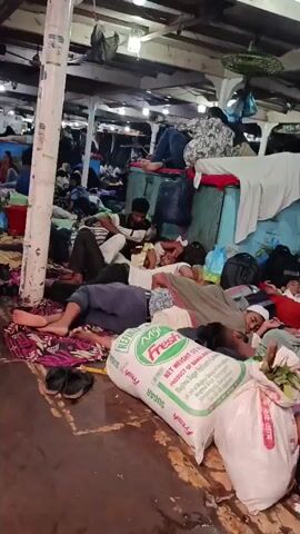 The video shows a large, open space crowded with people, mostly lying down on mats or blankets. Some are sleeping, while others are sitting and looking at their phones. There are large white bags stacked along one side of the room, and personal belongings like backpacks and clothing are scattered throughout the space. The room has a low ceiling with exposed beams and fluorescent lighting. There are also support pillars throughout the room. Some people are walking around, adding to the sense of a crowded and makeshift living space.