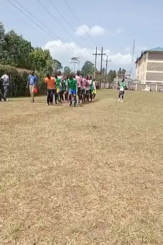The video shows a group of young people jumping on a grassy field, possibly warming up or performing a drill. The participants are wearing various athletic attire, including jerseys of different colors (green, pink, orange) and shorts. They are jumping in unison, moving forward across the field. There are some observers standing on the side, and a building is visible in the background. Power lines stretch across the sky, and trees line the edge of the field. The ground appears to be dry grass, indicating it might be a warm day.