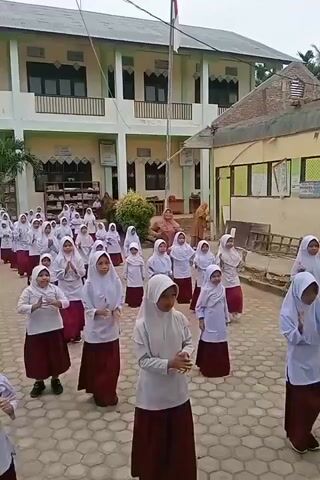 A group of young girls in white hijabs and maroon skirts are gathered outdoors, appearing to be in a schoolyard. They are standing in rows and performing synchronized movements, possibly a dance or exercise routine, with their hands extended and moving. The girls are facing a building with multiple stories, featuring a light green exterior with white trim, balconies, and numerous windows. A flagpole stands in front of the building. Two adult women, also wearing hijabs, are visible in the background, observing the group. The ground is paved with hexagonal tiles. The scene appears to be filmed during daylight, under an overcast sky.