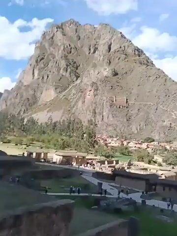 The video shows a panoramic view of a town nestled between two large, verdant mountains, partially covered with buildings and foliage.  The town has rows of houses with visible roofs.  The camera pans over the townscape, revealing people walking on the ground, some descending a stone structure. The landscape suggests a historic site or an ancient settlement.  The sky is blue, scattered with white clouds.  The overall setting evokes a sense of exploration and historical significance, with the mountainous backdrop enhancing the scene's grandeur.