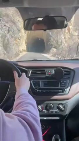 The video shows a driver's perspective inside a car as it approaches and drives through a tunnel. The car's dashboard, steering wheel, and rearview mirror are visible, with the driver's hands on the wheel. Outside the car, the tunnel is surrounded by rocky terrain. The car drives into the tunnel, briefly obstructing the view of the outside environment.