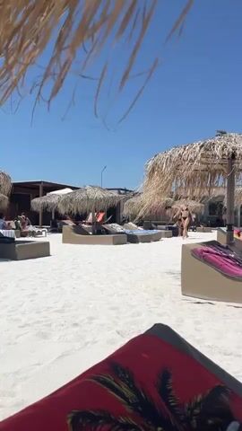 The video displays a poolside resort scene on a sunny day. The camera, positioned from a first-person perspective, captures a view of a white sandy beach with thatched umbrellas and lounge chairs. Several people are visible relaxing on loungers, some sunbathing, others reading or using their phones. In the background, a swimming pool is filled with people, and palm trees and buildings are visible on the horizon. The video shows close-ups of people in swimwear and their feet, emphasizing the relaxing atmosphere of the resort.