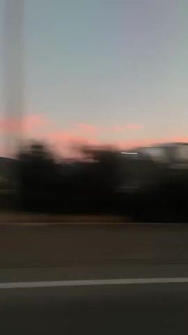The video shows a blurred view of a sunset over a dark treeline. The sky is a gradient of pale blue and pink. In the foreground, there is a gray road with a white line. The movement suggests the video was filmed from a moving vehicle.