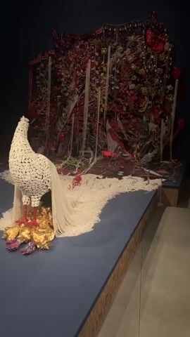 The video showcases an intricate art installation located in Venice, Italy. The central piece is a large, white, peacock-like sculpture positioned on a blue platform. Its body is composed of densely packed, small, white elements, while its expansive tail unfurls across the platform, made from countless white, bead-like strings that create a wavy, textured surface. The sculpture's feet are red and it stands on a cluster of gold, irregular, crumpled shapes. Behind the peacock, a large, dark background artwork is visible, adorned with rich textures, shiny red and green elements, and what appear to be various embellishments like beads or sequins. The camera pans and zooms, offering close-up views of the delicate details of the white tail and the vibrant textures of the background piece. No people or visible text are present in the video.