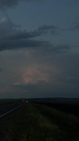 This is a vertical, eye-level video captured during dusk or twilight, as indicated by the dark, cloudy sky and the lit headlights of a vehicle. The scene depicts a rural highway in Kurgany, Russia, with a paved road stretching into the distance. To the right of the road is a grassy embankment and a dark field, possibly farmland. The sky is filled with dramatic, dark clouds, with a lighter, possibly illuminated patch of clouds in the middle distance, suggesting either distant lightning or an atmospheric phenomenon. A single truck with its headlights on is visible on the road, suggesting travel. The overall mood is somber and atmospheric, evoking a sense of isolation and the vastness of nature. There is no visible text.