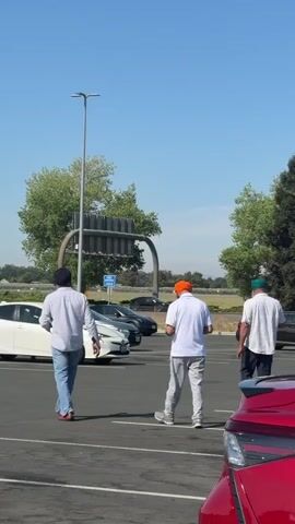 The video shows three men walking away from the camera in a parking lot. They are all wearing light-colored shirts and pants, and each has a colorful turban on their head: one orange, one blue, and one green. The men are walking towards a white car parked in the lot, with other cars visible in the background. The sky is clear and blue, and trees are visible in the distance. The camera appears to be positioned from within a red car, with the front corner of the red car visible in the lower right portion of the frame.