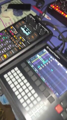 A person's hands are visible, interacting with a music production setup. The setup includes at least two electronic devices with buttons and screens, likely MIDI controllers or sequencers. One device, on the left, has colorful, illuminated buttons and a small screen displaying text and graphics. Another device, in the foreground, features a grid of white buttons and a larger screen showing a digital audio workstation interface with multiple tracks and waveforms. A third device, on the right, is partially visible and appears to be a rackmount audio unit with red illuminated buttons. A guitar effects pedal, labeled "Strymon NIGHTSKY," is also visible in the lower right corner. The background suggests an indoor or outdoor setting with a blue and white patterned fabric underneath the equipment and a glimpse of what might be grass or dirt. The person's hands are actively pressing buttons and manipulating controls on the devices, indicating they are in the process of creating or performing music. The person is wearing a shirt with a leopard print pattern on the sleeve. There are several cables connected to the equipment.