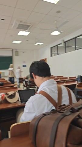 The video shows the interior of a classroom.  People are seated at desks, some are looking towards the front of the room where a presentation is being displayed on a projector screen.  There is a chalkboard behind the presenter's area.  Large windows line one side of the room. Some people are walking around. The camera moves through the classroom and focuses on the wooden desks. The last shot is of the inside of a desk drawer.