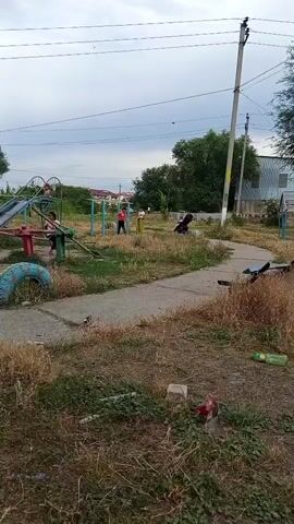 This video shows a playground in Belovodskoe, Kyrgyzstan. The playground has several slides, a seesaw, and other climbing structures, but appears to be in a state of disrepair. The ground is covered in dry grass and debris, and some of the playground equipment is rusted or broken.

Several people, mostly children, are present on the playground. One child is on a slide, another is on the seesaw, and a few others are walking around. The weather appears to be overcast, and the time of day is likely afternoon, given the light. The overall impression is one of a neglected community space. There is no visible text in the video.
