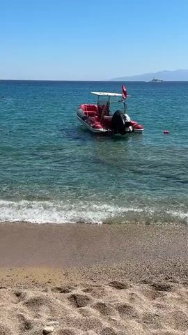 A rigid-hulled inflatable boat (RHIB) is anchored in clear, blue water just offshore from a sandy beach. The boat is red and white with a black outboard motor and a canopy. A person, wearing a red top and dark shorts, is visible seated in the boat. A red buoy floats nearby. In the distance, a faint landmass with a structure on it is visible under a clear blue sky. Gentle waves are lapping at the sandy shore in the foreground.