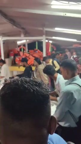The video shows a crowded ferry with many people and motorcycles. The ferry appears to be at night or in low light conditions, illuminated by overhead fluorescent lights. People are standing and sitting, some near motorcycles. Life jackets are visible stacked in the background. The atmosphere seems busy and somewhat chaotic as people and vehicles are packed together on the ferry.
