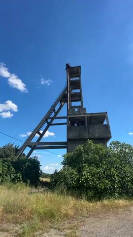 The video shows a time-lapse of an abandoned concrete mine headframe structure against a clear blue sky with scattered clouds. The structure is tall and appears to be a series of interconnected concrete platforms and beams, with a steep incline leading to the top. There is dense green foliage and dry grass in the foreground. The camera remains stationary throughout the recording.