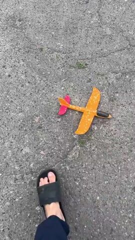 The video begins with a close-up of a toy airplane with an orange body and pink and orange wings resting on a grey asphalt surface. The airplane is positioned diagonally from the bottom right to the top left of the frame. Small green weeds are visible growing through cracks in the asphalt. The camera then pans to the right, revealing more of the asphalt surface and a small patch of grass. As the camera continues to move, the scene transitions to an outdoor setting. A narrow, paved road stretches into the distance, flanked by a grassy verge and trees on the left. On the right side of the road, there are several low-lying buildings, some appearing dilapidated, with corrugated metal roofs and grey walls. A wooden fence and more vegetation are visible. Further down the road, utility poles with wires are present. The sky is overcast with grey clouds. The video appears to be taken from a moving perspective, possibly while someone is walking or driving slowly.