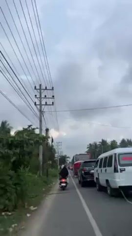 The video depicts a road with heavy traffic, primarily consisting of buses, cars, and a single scooter. The scooter, driven by a person wearing a head covering and jacket, is moving in between the vehicles. The vehicles are stopped or moving very slowly. The sky is overcast and a line of power lines run along the roadside near some trees. The overall scene suggests a congested roadway in a suburban or rural area.