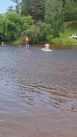 The video shows a wide river with several people on paddleboards. In the foreground, the water is clear and shallow, with pebbles visible on the riverbed. In the middle ground, four individuals are paddleboarding. One person, wearing an orange life jacket, is on a blue paddleboard in the center. To the right of them, another person is on a yellow paddleboard. To the left, two more people are on red paddleboards, further upriver. The background consists of a lush, green treeline with mature trees and foliage. The sky above is bright, suggesting a sunny day. The water appears calm with gentle ripples.