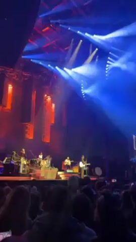 A band is performing on stage in front of a large crowd at an outdoor venue. There are seven musicians on stage playing various instruments including drums, guitar, and keyboard. The stage is illuminated with bright red and blue lights that are shining on the performers. The crowd is dense and consists of people of different ethnicities, many of whom are holding up their phones to record the performance. The venue appears to be a large open space with a structured ceiling visible above the stage lights.