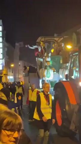 A night-time street scene displays a parade. A large, orange tractor tows a decorated float. The float has a green, horse-drawn carriage theme with people dressed in costumes. A person in a red shirt and gold pants stands on the float and interacts with the crowd. Several people, wearing yellow safety vests, walk alongside the float. Many other people are watching from the side of the street. The scene is illuminated by streetlights and lights on the buildings.